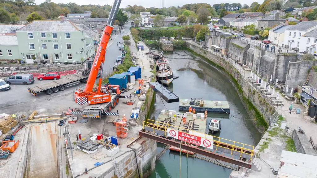 A drone view looking from the seaward end of Charlestown Harbour over the lock gate into the inner harbour. A crane on the dockside is lifing the first section of the new lock gate onto a pontoon from a flat bed lorry.