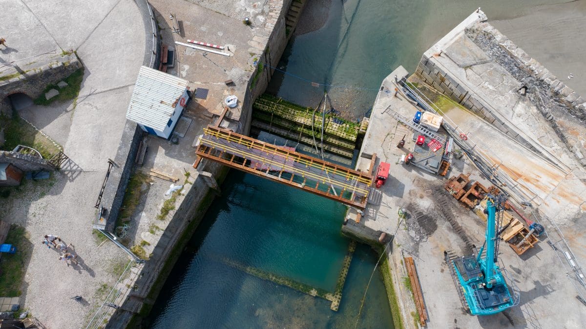 An aerial photograph of the lock gate at Charlestown Harbour looking straight down from above. There is a temporary bridge and wave attenuation blocks where the old lock gate once stood. There is a crane and other signs of work on the quayside.