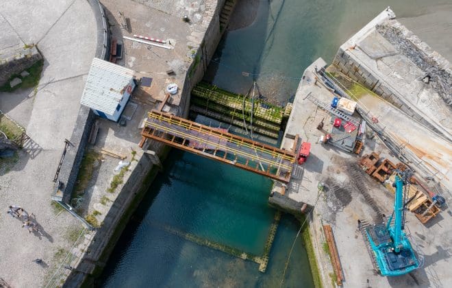 An aerial photograph of the lock gate at Charlestown Harbour looking straight down from above. There is a temporary bridge and wave attenuation blocks where the old lock gate once stood. There is a crane and other signs of work on the quayside.
