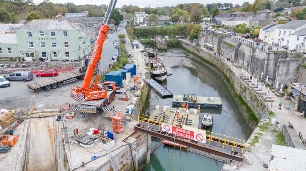 A drone view looking from the seaward end of Charlestown Harbour over the lock gate into the inner harbour. A crane on the dockside is lifing the first section of the new lock gate onto a pontoon from a flat bed lorry. 